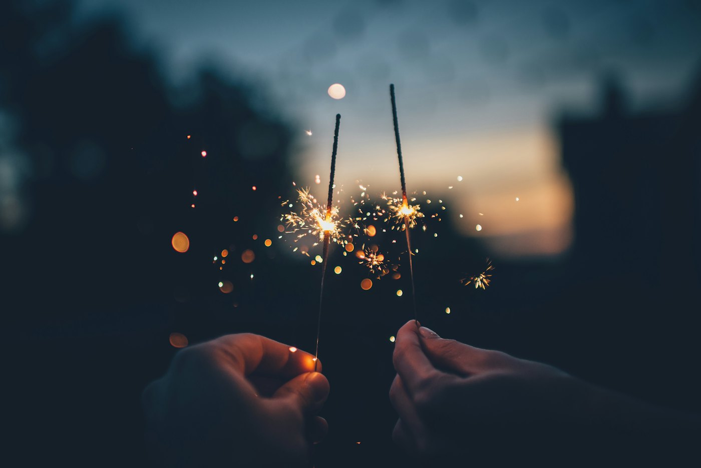 Two hands holding lit sparklers against an evening sky